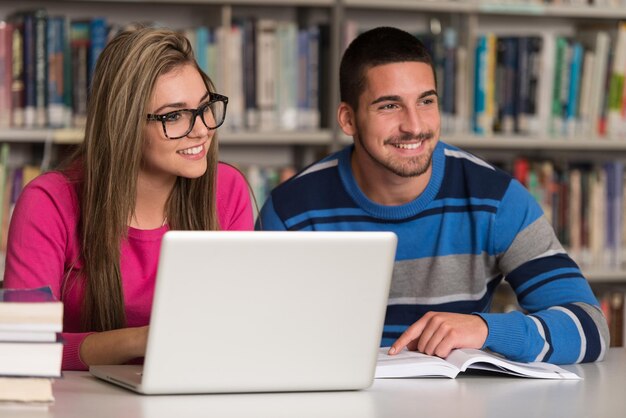 En la biblioteca Dos estudiantes universitarios guapos con computadora portátil y libros que trabajan en una biblioteca universitaria de secundaria con poca profundidad de campo