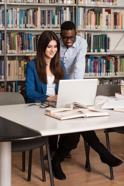 En la biblioteca Dos estudiantes universitarios guapos con computadora portátil y libros que trabajan en una biblioteca universitaria de secundaria con poca profundidad de campo