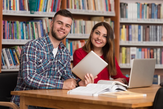 En la biblioteca Dos estudiantes universitarios guapos con computadora portátil y libros que trabajan en una biblioteca universitaria de secundaria con poca profundidad de campo