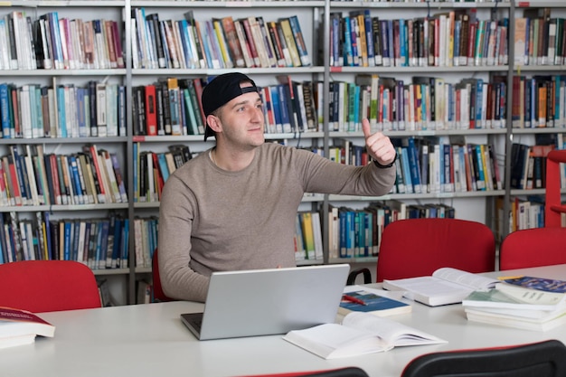 En la biblioteca Estudiante masculino con gorra trabajando en una computadora portátil y libros en una biblioteca universitaria de secundaria