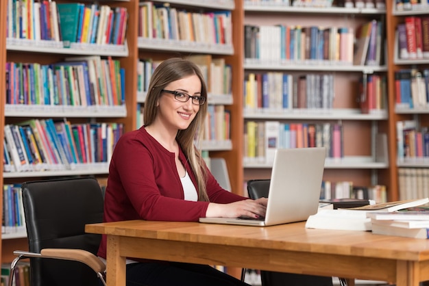 En la biblioteca Hermosa estudiante femenina con computadora portátil y libros que trabajan en una biblioteca universitaria de secundaria con poca profundidad de campo