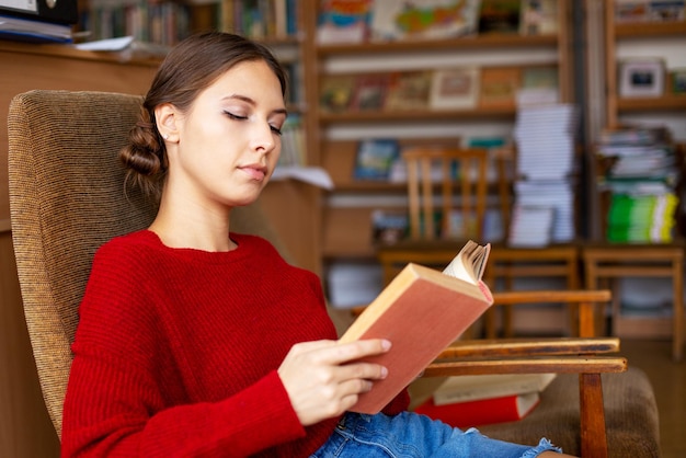 Biblioteca universitária, linda garota caucasiana, sentada na cadeira ao lado da estante, segurando o livro e ...