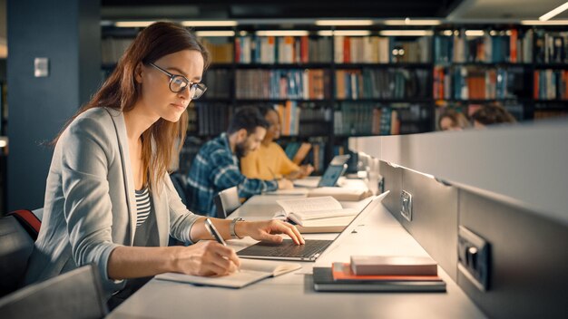 Biblioteca universitaria Niña caucásica talentosa sentada en el escritorio utiliza la computadora portátil escribe notas para el ensayo de papel Estudio para la asignación de clase Grupo diverso de estudiantes Aprendiendo Estudiando para exámenes