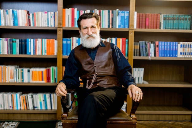 bibliotecaria del profesor académico, vestido con camisa y pantalones oscuros y chaleco de cuero, sentado en una silla en la biblioteca, bibliotecas en el fondo. Concepto de conocimiento, aprendizaje y educación.