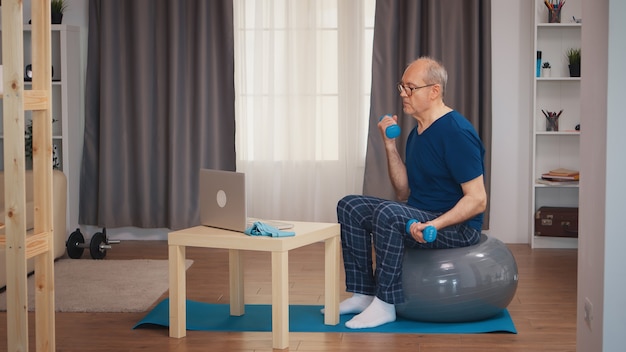 Bíceps de entrenamiento de hombre mayor viendo la lección de fitness en línea. Anciano pensionista entrenamiento saludable deporte sanitario en casa, ejercicio de actividad física en la vejez