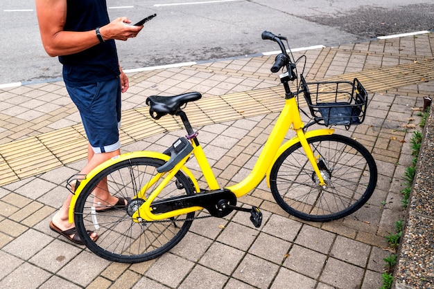 BICICLETA AMARILLA y teléfono móvil. Hombre que usa el servicio de alquiler de vehículos e con teléfono inteligente en la calle y el parque urbanos de la ciudad. Joven alquila un transporte ecológico en verano.