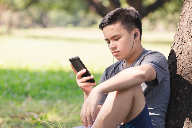 Bicicleta asiática joven que hace una pausa árbol y que escucha la música con el auricular