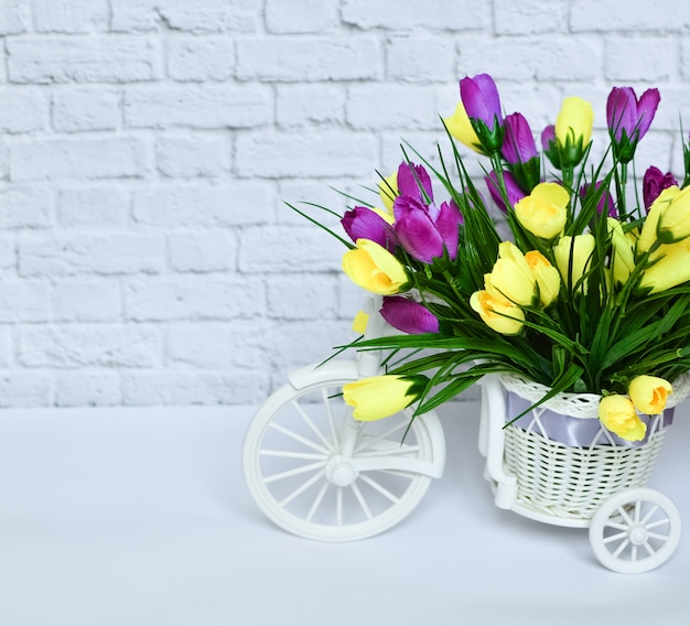 Bicicleta decorativa bonita pequena com as flores amarelas e roxas em um fundo branco.