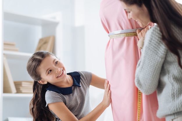 Esta bien. Feliz niña positiva positiva tocando un modelo de vestido y mirando a su madre mientras espera una aprobación