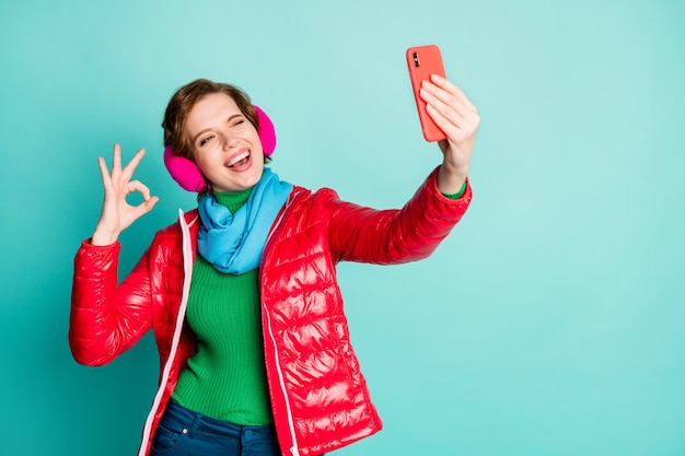 ¡Bien! Foto de señora bonita divertida mantenga el teléfono haciendo selfies guiñando el ojo mostrando bien símbolo usar bufanda de abrigo rojo orejeras rosadas pantalones de puente aislado pared de color verde azulado