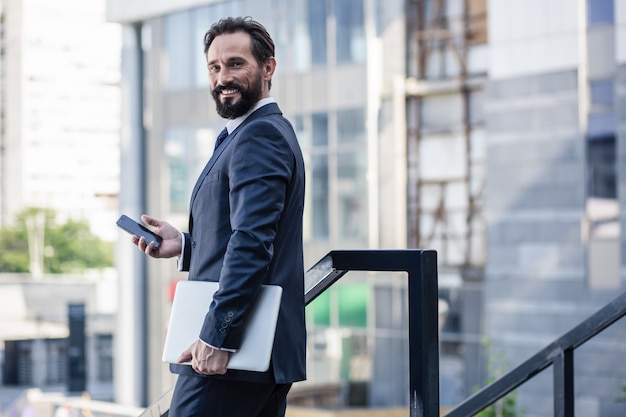 Bien vestido. Cintura para arriba de un empresario barbudo positivo sosteniendo su computadora portátil mientras usa el teléfono inteligente