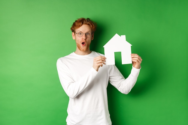 Bienes raíces y concepto de compra de propiedad. Hombre joven pelirrojo sorprendido mostrando el modelo de la casa de papel y mirando asombrado, de pie sobre fondo verde.