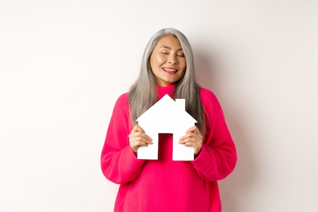 Bienes raíces. Hermosa dama asiática soñadora abrazando a modelo de casa de papel con los ojos cerrados, sonriendo como soñando con comprar un apartamento, de pie sobre fondo blanco.