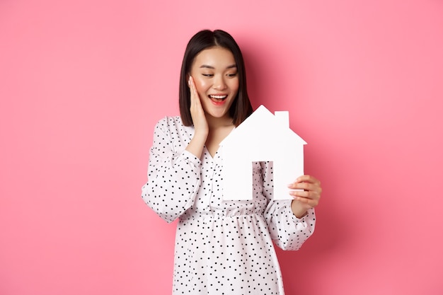 Bienes raíces. Linda mujer asiática en busca de apartamento, mirando feliz al modelo de casa de papel, de pie contra rosa