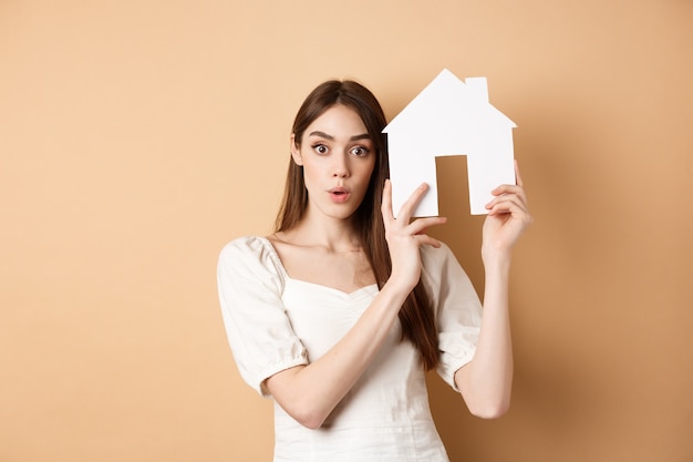 Bienes raíces. Mujer joven emocionada que muestra el recorte de la casa y mirando a la cámara, alquilando una propiedad, de pie en beige.