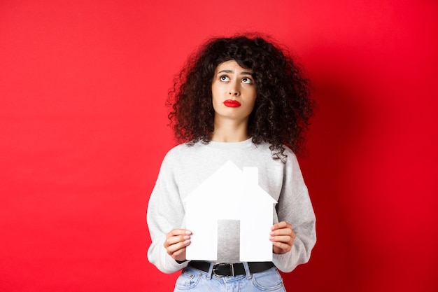 Bienes raíces. Mujer triste soñando con comprar un apartamento, sosteniendo un recorte de la casa de papel y mirando hacia arriba angustiado, de pie en la pared roja.