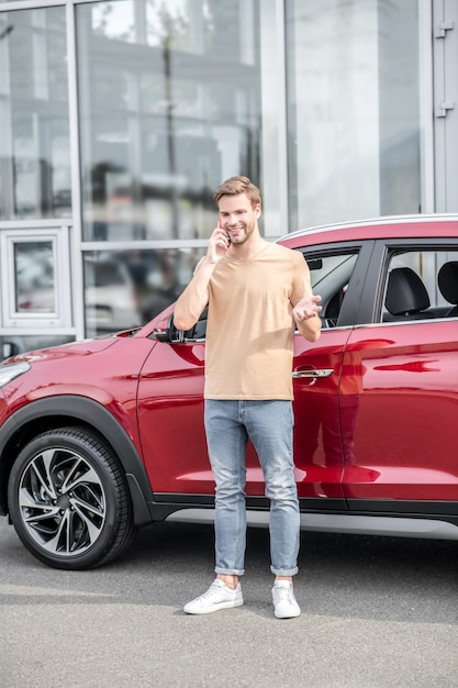 Bienestar. Hombre adulto joven feliz en ropa casual divirtiéndose hablando por teléfono inteligente mientras gesticula cerca del coche rojo en el fondo del edificio