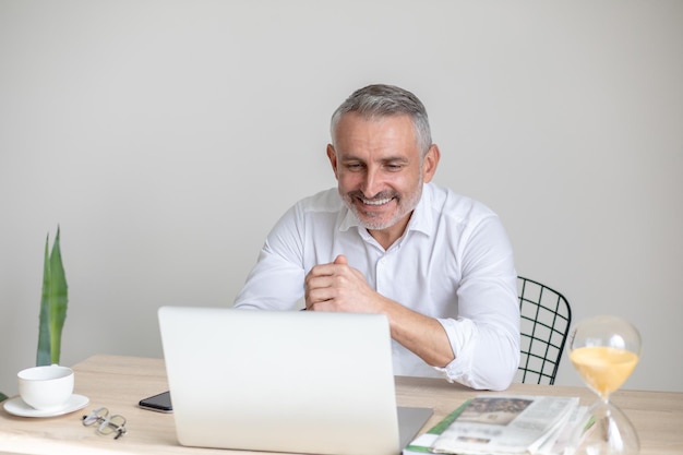 Bienestar. Hombre canoso feliz con camisa blanca mirando a la computadora portátil mientras se sienta en el lugar de trabajo en la mesa en una habitación luminosa