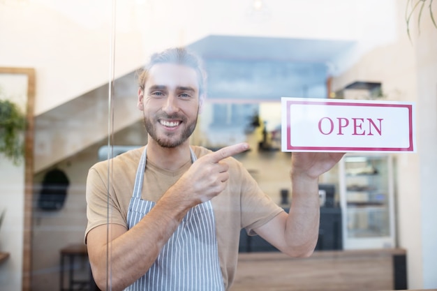 Bienvenida. Feliz joven barbudo en delantal de pie detrás de un vidrio dentro del café apuntando al cartel que dice Abrir en la entrada