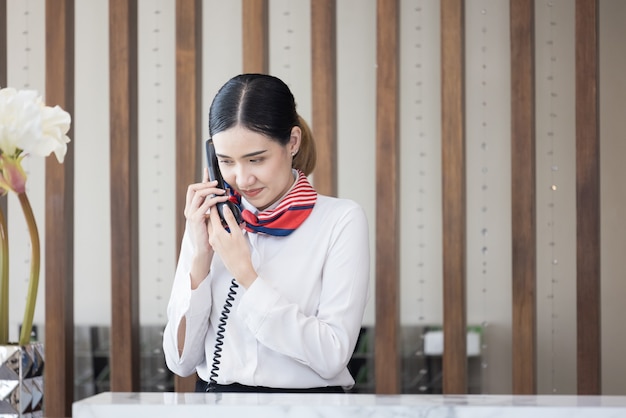 Bienvenido al hotel, feliz joven asiática trabajadora recepcionista de hotel sonriendo de pie, tomando una llamada telefónica en un mostrador de recepción de lujo moderno esperando a que los huéspedes obtengan la tarjeta de acceso en el hotel