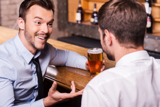 Bier mit einem guten Freund teilen. Zwei fröhliche junge Männer in Hemd und Krawatte, die miteinander reden und gestikulieren, während sie an der Bartheke Bier trinken