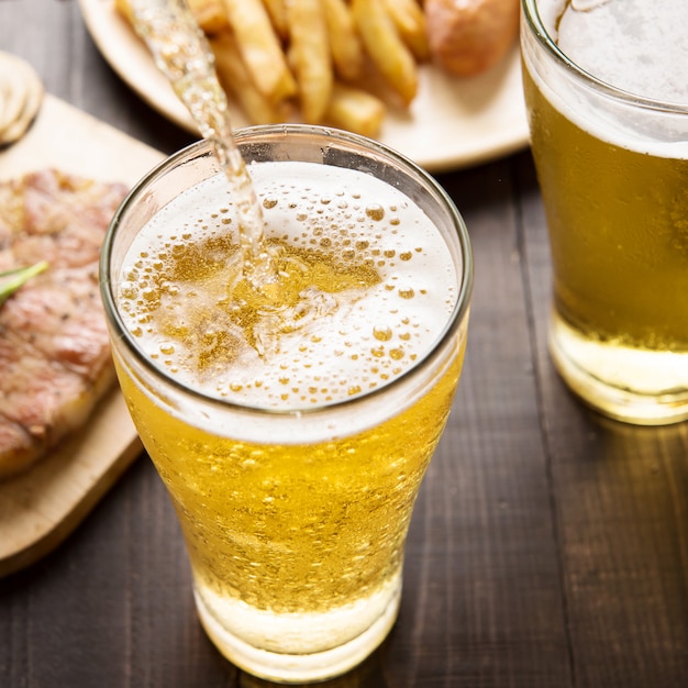 Bier wird in Glas mit Steak und Pommes Frites auf hölzernem Hintergrund gegossen