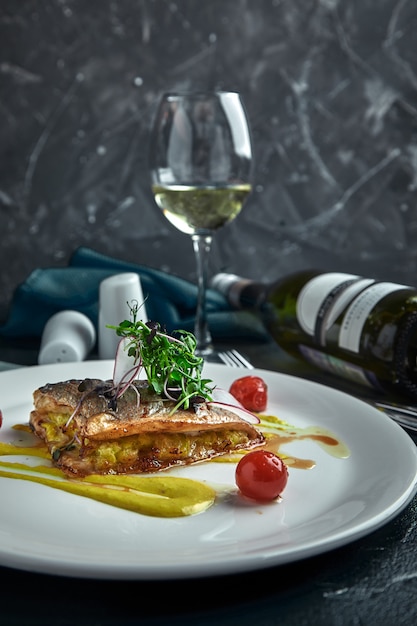 Bife branco de peixe grelhado no travesseiro de legumes e molho amarelo, com tomates e ervas frescas. Pesque no prato branco com vinho branco na parede escura bonita. Fechar-se. Espaço