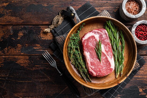 Bife de costilla cruda, carne de buey de costilla veteada en placa de madera con romero y tomillo. Fondo de madera. Vista superior. Copie el espacio.