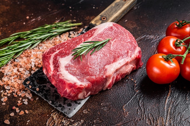 Bife de costilla cruda sin cocer, carne de ternera chuletón en una cuchilla de carnicero con hierbas. Fondo oscuro. Vista superior.