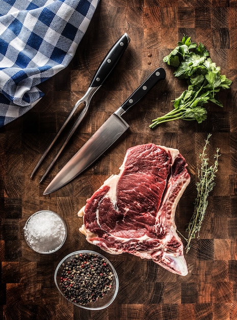 Bife de costilla con hueso sobre tablero de carnicero con hierbas sal pimienta tenedor y cuchillo.