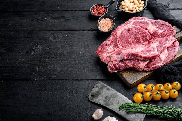 Bife cru, pimenta, ervas, alho, alecrim do conjunto de carne bovina orgânica em mármore e uma velha faca de cutelo de açougueiro, no fundo da mesa de madeira preta, com espaço de cópia para o texto