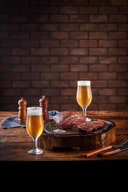 Bife de alcatra grelhado cap na tábua de madeira com dois copos de cerveja de tulipa fria suado. Mesa de madeira e fundo de parede de tijolos (picanha brasileira).