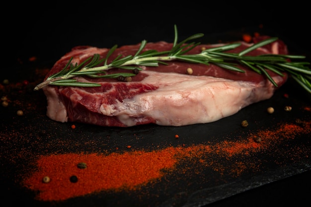 Bife de bife marmorizado cru com ramo de alecrim e especiarias. Um pedaço de carne com veias gordurosas em uma placa preta de pedra. Vista lateral.