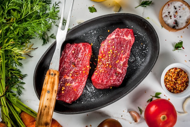 Bife de carne Bifes crus frescos em uma frigideira Especiarias (pimenta salgada) legumes frescos - tomates cenouras cebolas alho Em uma mesa de mármore branco com um garfo para carne e uma faca