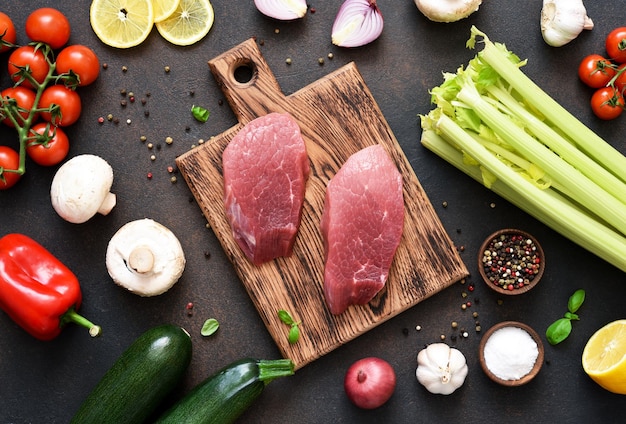 Bife de carne crua em uma placa com vegetais diferentes em um fundo de concreto.