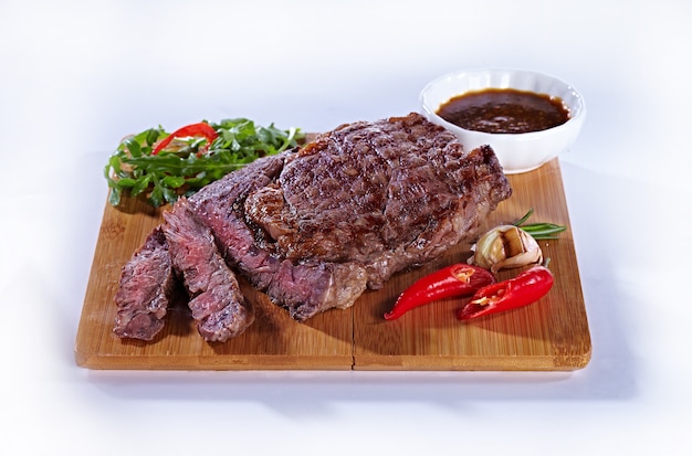 Bife de carne frito delicioso suculento médio sobre uma tábua de madeira, com pimenta, alho, ervas e molho de tomate. Bife para um restaurante em um fundo branco