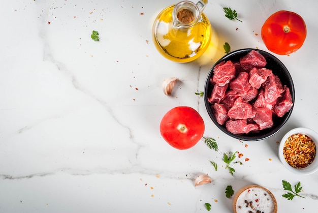 Bife de carne. Goulash picado cru fresco, cubos de carne em uma tigela. Especiarias (sal, pimenta), tomate, alho, cebola. Em uma mesa de mármore branco, com um garfo para carne e uma faca. vista do topo