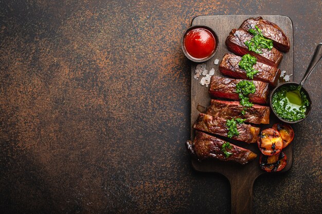 Bife de carne marmorizada grelhada / frita e fatiada com garfo, tomate, molhos diferentes na tábua de madeira, vista superior, close-up com espaço para texto, fundo de pedra rústico. Conceito de bife de carne bovina