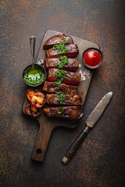 Bife de carne marmorizada grelhada ou frita e fatiada com garfo, tomate como acompanhamento e diferentes molhos na tábua de madeira, vista superior, close-up, fundo rústico de pedra. Conceito de bife de carne bovina