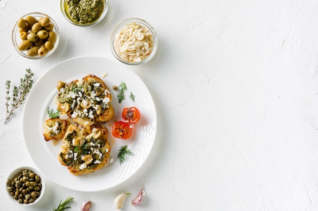 Bife de couve-flor com especiarias molho chimichurri flocos de amêndoa azeitonas tomate cereja frito e alcaparras em um prato branco Comida vegetariana Fundo branco Copyspace