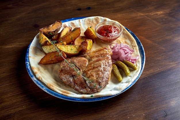 Bife de porco grelhado com temperos e guarnição de batata, servido em um prato com cebolas em conserva, pepino e molho vermelho sobre uma superfície de madeira