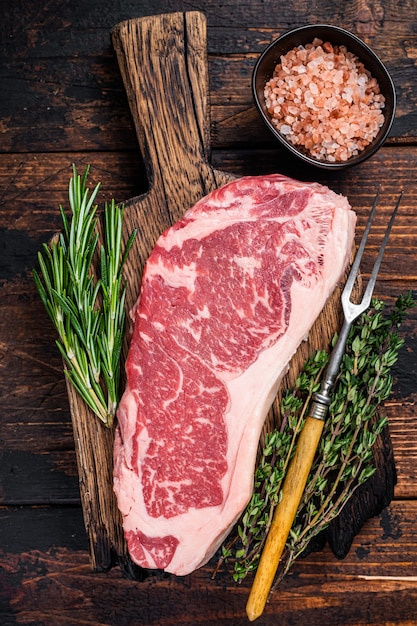 Bife de tira de Nova York crua ou striploin em uma placa de madeira. Fundo de madeira escuro. Vista do topo.