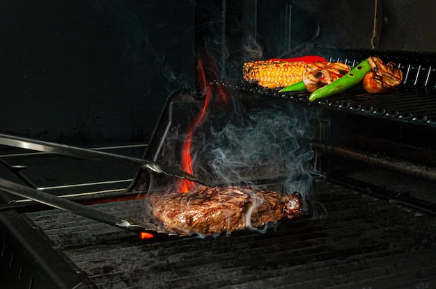 Bife e legumes são grelhados e virados com pinças. a foto mostra chamas e fumaça. fechar-se.