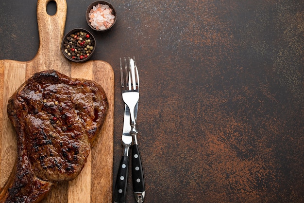 Bife grelhado ou frito com carne nobre em mármore Ribeye servido em tábua de madeira com talheres. Vista superior de um suculento bife cozido em um fundo rústico de concreto marrom com espaço para seu texto