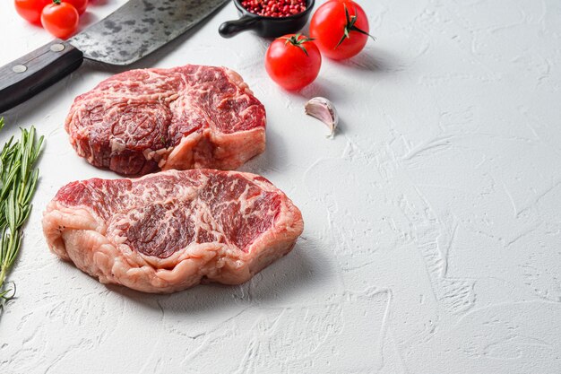 Bife orgânico, carne bovina crua com temperos, alecrim e cutelo de açougueiro. Mesa texturizada branca. Vista lateral.