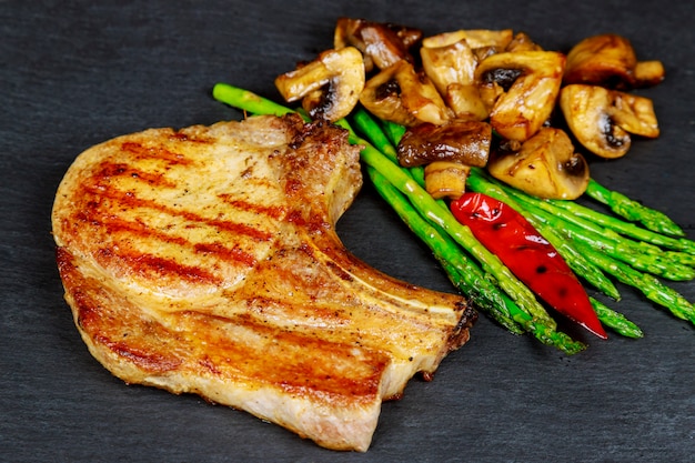 Bifes de carne de porco grelhada com ervas, aspargos, cogumelos em fundo de ardósia de pedra