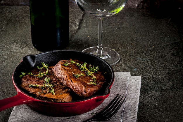 Bifes de carne grelhados caseiros em uma frigideira com porções, com um garfo, faca e um copo de vinho na mesa de pedra preta