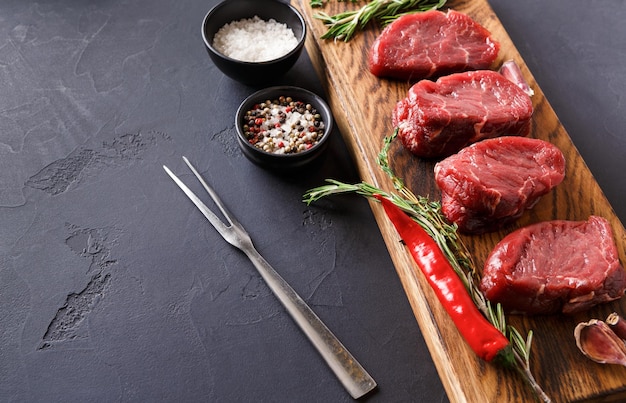 Bifes de filé mignon crus com ervas e especiarias. Cozinhar ingredientes para prato de restaurante. Carne fresca, sal, alecrim, alho e pimenta na placa de madeira, garfo de carne em fundo preto com espaço de cópia