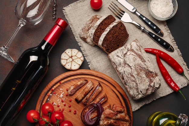 Bifes de porco grelhado fatiado com garrafa de vinho, copo de vinho, saca-rolhas, faca, garfo, pão preto, tomate cereja, alho