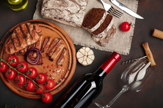 Bifes de porco grelhado fatiado com garrafa de vinho, copo de vinho, saca-rolhas, faca, garfo, pão preto, tomate cereja, alho